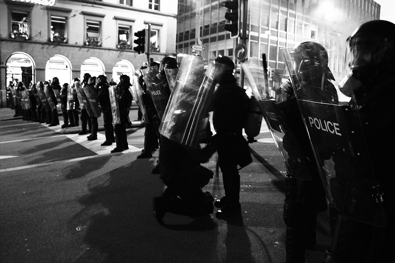 Policeman in riot control gear checking equipment of another policeman.
