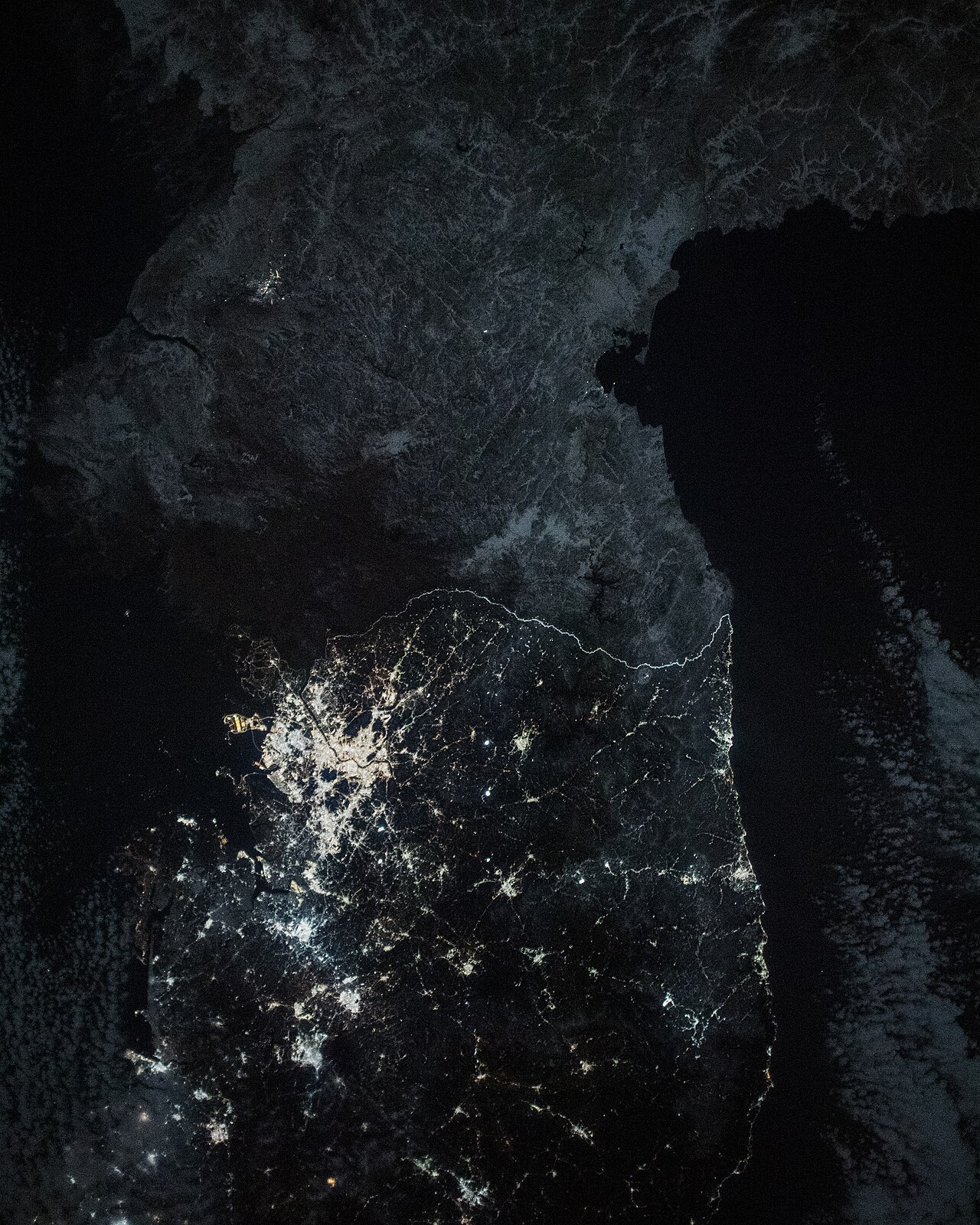 Nighttime satellite image of the Korean Peninsula with North Korea appearing dark and South Korea lit with city lights.